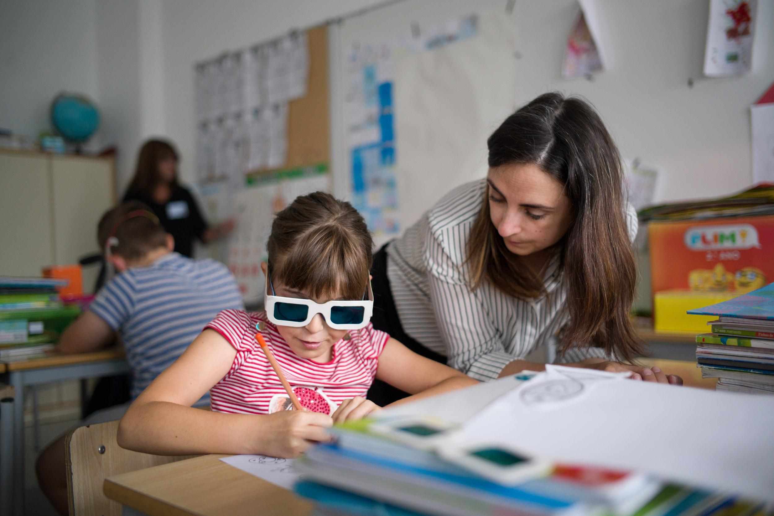 Adult with long dark brown hair next to a child with ColorADD glasses on (helps colorblind kids see color clearly). They are working on the child's writing