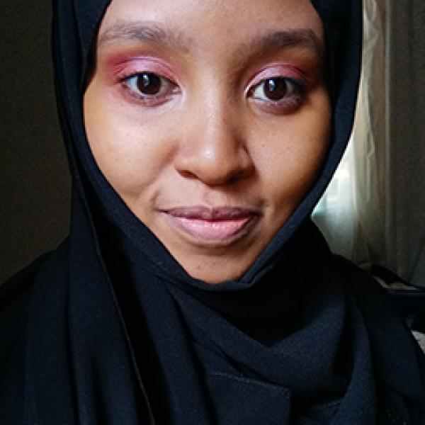 Black woman, wearing a black hijab. She is slightly smiling and wearing a light pink eyeshadow. 