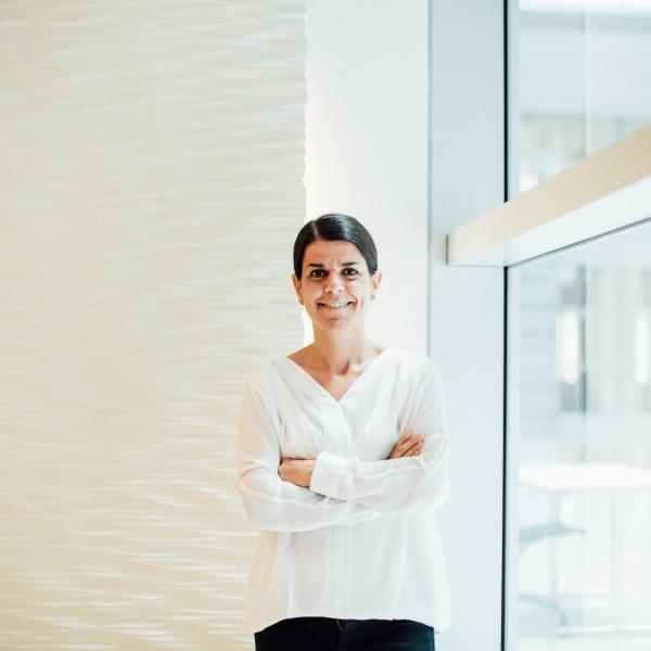 Photo of Ana Saenz de Miera; person dressed in a white shirt smiling at the camera, arms crossed. Background of a corner in between a white wall and a large window overlooking a street
