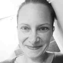 Black and white photo of Kristina Schraitel of the Ashoka UK office. Person with lighter skin, brown hair and a green top smiling at the camera. Background a flight of stairs