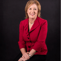 Photo of Leadership Group Member Lisa Davis. Person with shoulder light red / dark blonde hair smiling at the camera. Dressed in a professional red blouse and black pants. Background of black