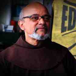 A black man, bald, with a beard and white hair, wearing a brown cassock. 