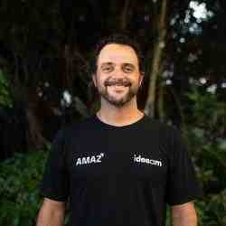 A white man wearing a black t-shirt in which is written "AMAZ" and "Idesam". He has short curly hair, moustache and short beard, with a few white hairs in his beard. He is smiling and his background there are some trees and plants, but they are a little blurred, as the camera focused on Mariano.