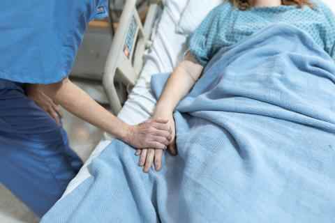 The photo shows a patient lying in a hospital bed under a blue blanket, a medical proffesional is holding the patient's hand and leaning over the bed. Their heads are not shown.