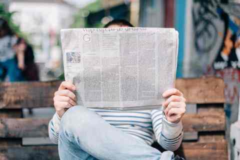 The photo shows a person sitting on a bench reading a newspaper. Their face is cover by the newspaper.