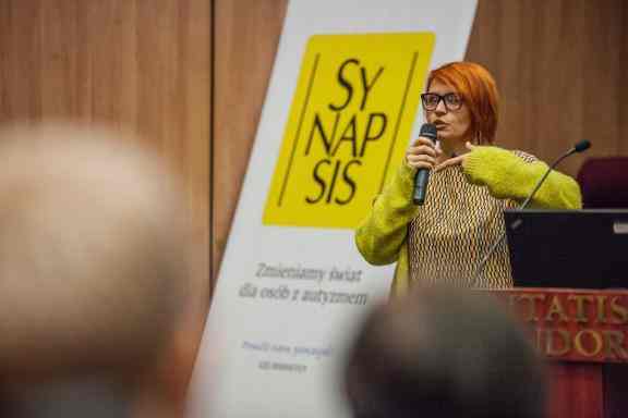Kobieta z krótkimi rudymi włosami mówi do mikrofonu na konferencji. W tle żółte logo fundacji Synapsis.