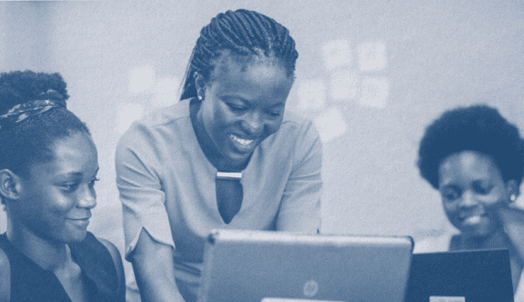 Photo shaded in dark blue. Three people huddle over a few laptops, smiling and looking at the laptops, as if they are working on something together