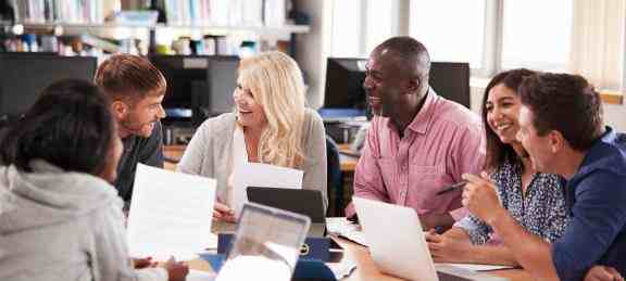 Grupo de pessoas estão conversando e sorrindo em torno de uma mesa em uma biblioteca. Algumas seguram folhas de papel, outras seguram canetas. Há tablets e computadores em cima da mesa