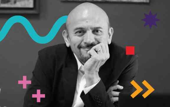 In the photo we can see Mauro - a Latin man, bald, wearing a suit and dress shirt. The photo is in black and white, and we have colorful graphic features added to the photo: a blue zigzag curve, a red square, a purple 8-pointed star, green dots, two pink plus signs and two orange arrows. 