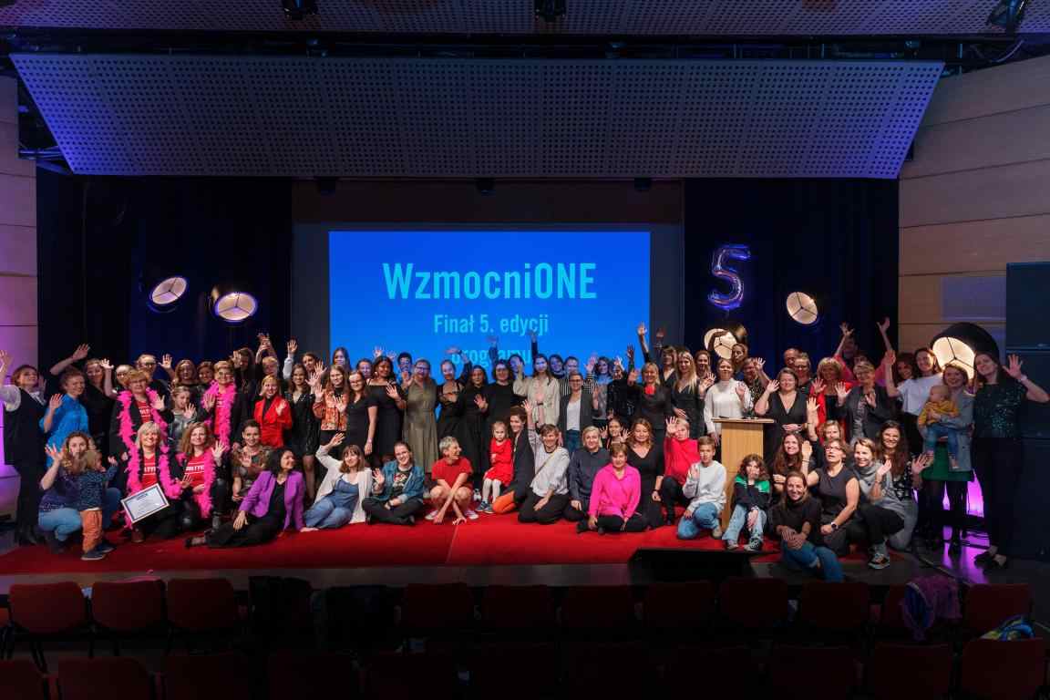 Zdjęcie przedstawia wszystkie osoby obecne na finale 5 edycji programu Wzmocnione na scenie w Muzem Manggha w Krakowie. Osoby uśmiechają się i machają, a za nimi widać rzutnimk z napisem "WzmocniONE. Finał 5 edycji."