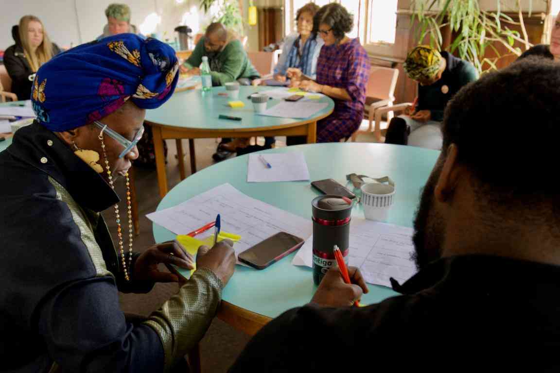 Group of adults speaking at a table together in a large room; background of other tables with groups of adults speaking