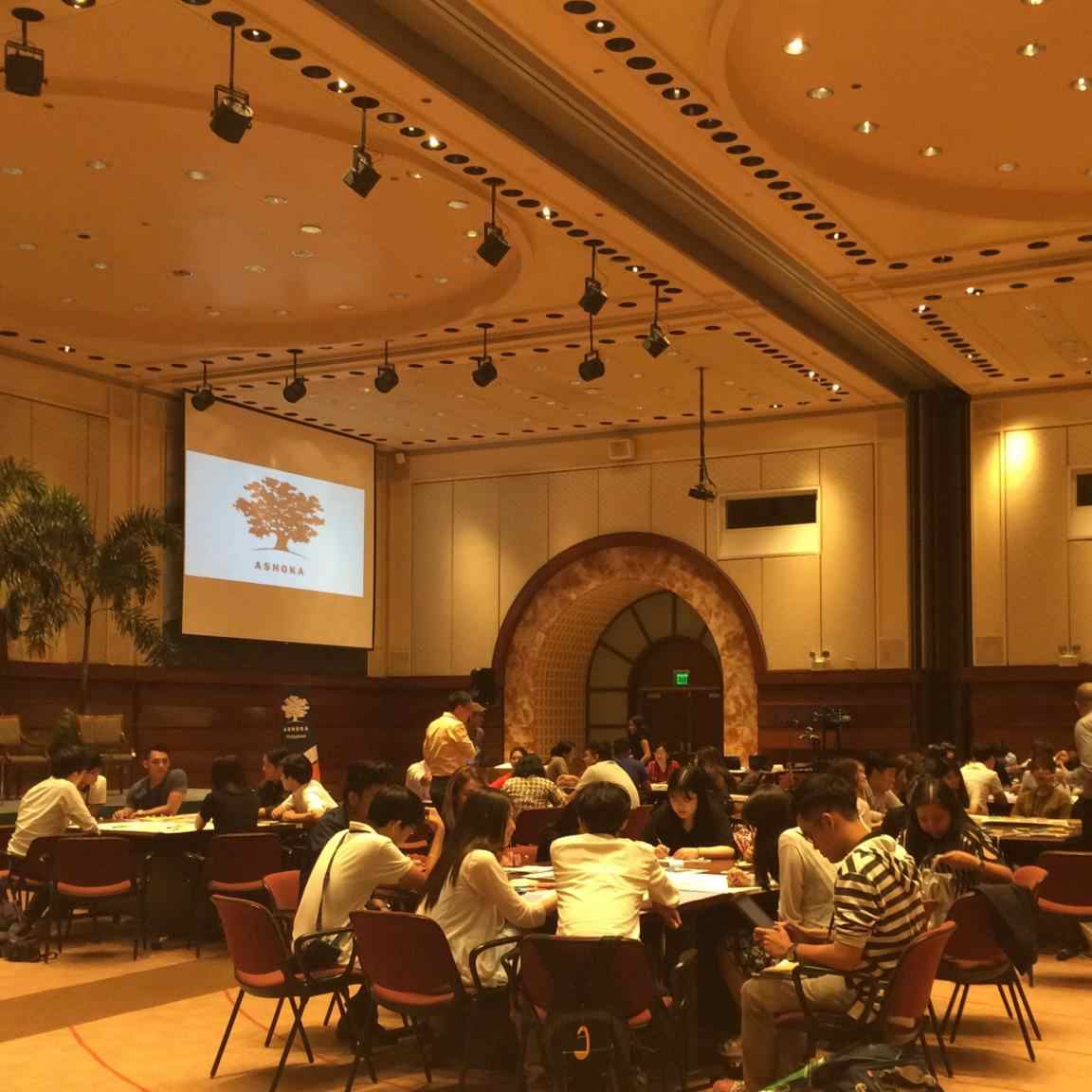 People arranged in circles around several tables in a warmly lit room with a high ceiling. In the background, the Ashoka logo is projected on a screen.