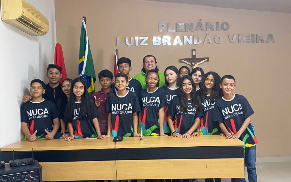 Vê-se uma foto de diversos estudantes com o Vereador Rodolfo Izodoro (Homem branco de cabelos longos e pretos) atrás de uma mesa com o escrito na parede atrás deles "Plenário Luiz Brandão Vieira"