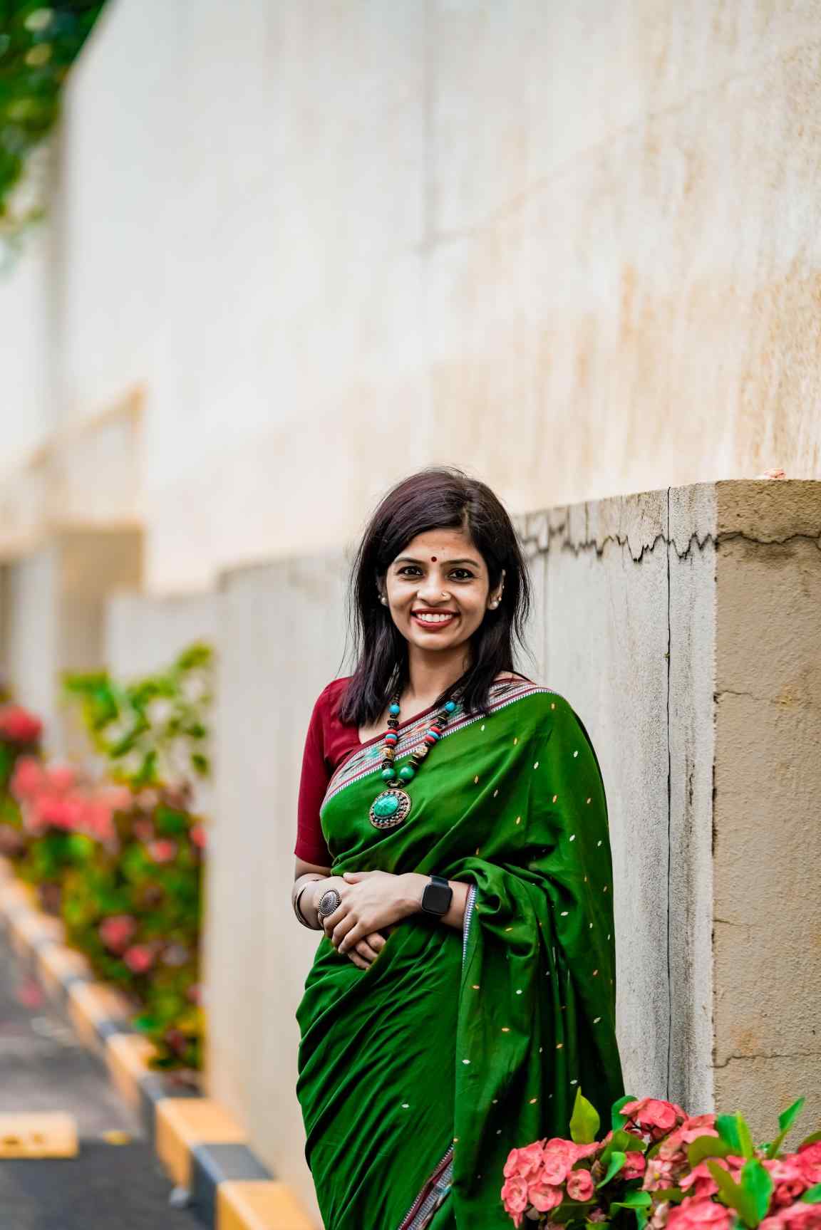 A headshot photo of Shruti Nair posing outside, in front of a stone wall with flowers