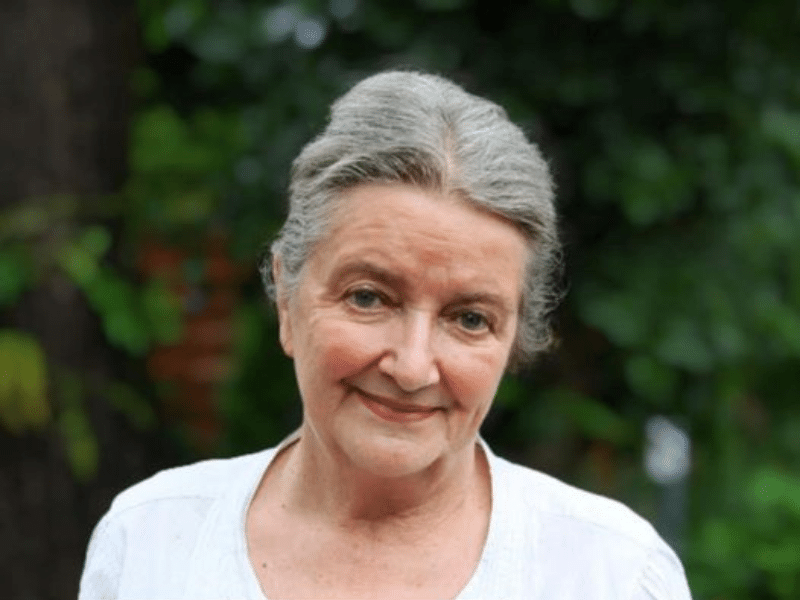 An older woman standing outside wearing white shirt, green trees in a background