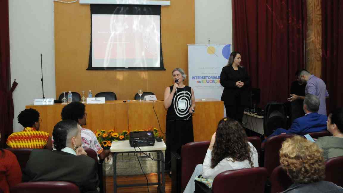 Vê-se Helena Singer (mulher branca de cabelos médios, lisos e grisalhos) segurando um microfone em frente a uma mesa em cima de um palco com diversas pessoas sentadas em cadeiras de frente para ela