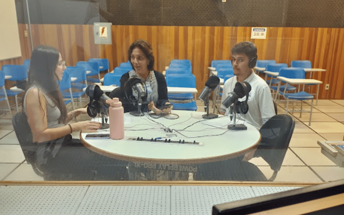 Três pessoas estão em um estúdio de gravação. Elas se sentam diante de uma mesa redonda de frente a microfones, e usam fones. Em primeiro plano, há um computador e uma mesa de áudio. Um vidro parece separar o grupo da mesa de áudio