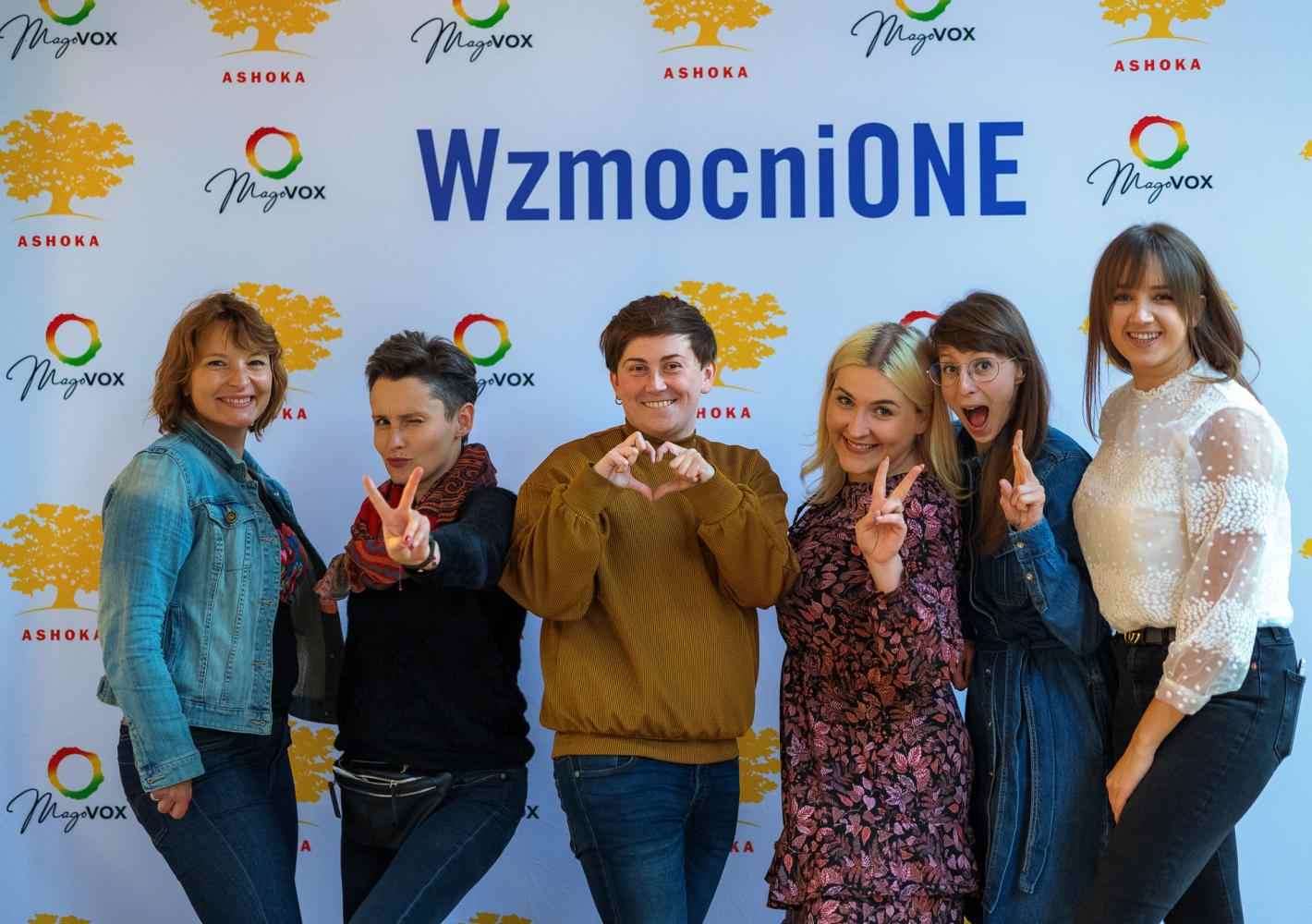 Na tle ścianki wzmocnionej z logo Wzmocnione, Ashoki oraz Magovox pozuje 6 osób w kolorowych ubraniach. Uśmiechają się, 3 pokazują znak peac/wolności palcami, jedna robi serduszko dłońmi.