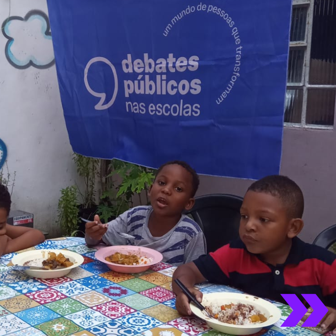Há duas crianças sentadas em uma mesa com seus respectivos pratos de comida, se alimentando. Logo atrás deles, há uma bandeira roxa dos Debates Públicos nas Escolas. Em destaque, lê-se "Debates Públicos nas Escolas", 