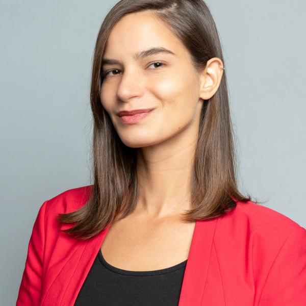 Headshot of Ashoka Fellow in Austria Rebekka Dober. Person with shoulder length brown hair smiling at the camera. Dressed in a bright red jacket and dark blouse. Background a white wall