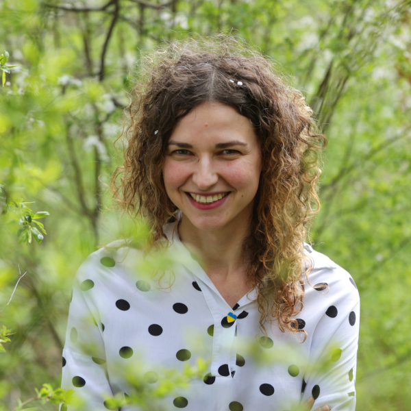 Young woman in a white shirt with black polka dots and with currly hair standing outside among the trees, smiling