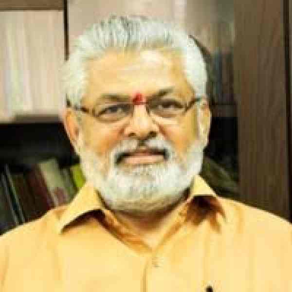 Photo of GG Gangadharan, Ashoka Fellow in India. Person with grey hair with glasses and a beard and dressed in a bright orange collared shirt smiling at the camera. There's an office in the background.