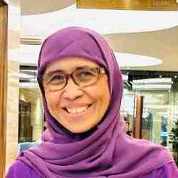 Person with purple head covering, purple top, and glasses smiling at the camera; background is a hallway