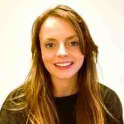 Person with shoulder length light brown hair and a dark sweater smiling at the camera. Background is a white wall