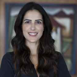 A white woman with wavy black hair with brown streaks, below shoulder height. She is smiling in the photo and wears a black shirt, a thin necklace with an orange pendant. Behind her we can see a painting, but the background is blurred 