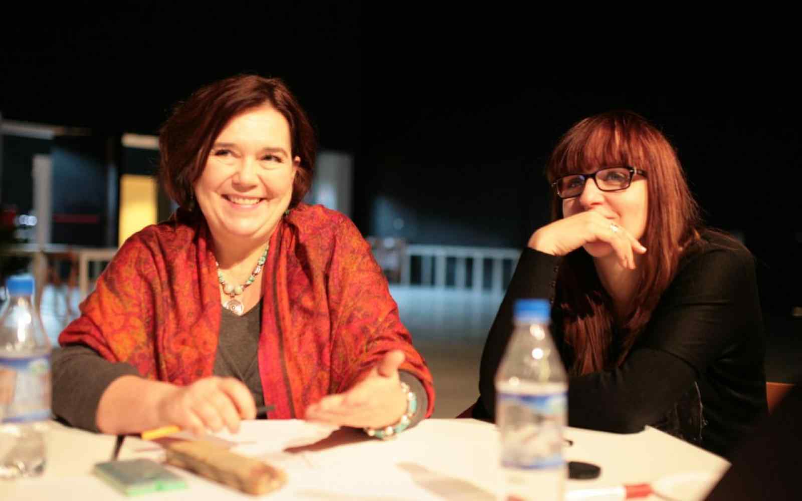 Zdjęcie przedstawia dwie uśmiechnięte kobiety siedzące przy stole. Po lewej Katarzyna Oleś w czerwonej chuście na ramionach i kolorowym naszyjniku.