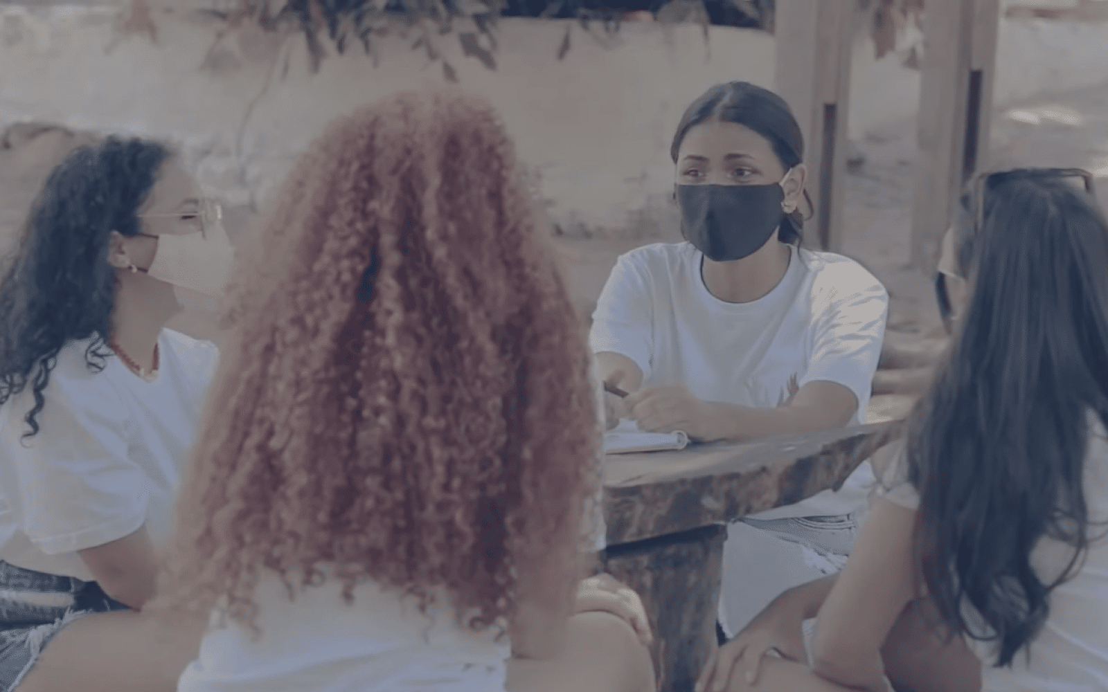 Grupo de 4 jovens mulheres conversando ao redor de uma mesa ao ar livre. Elas usam camiseta branca e máscara de proteção