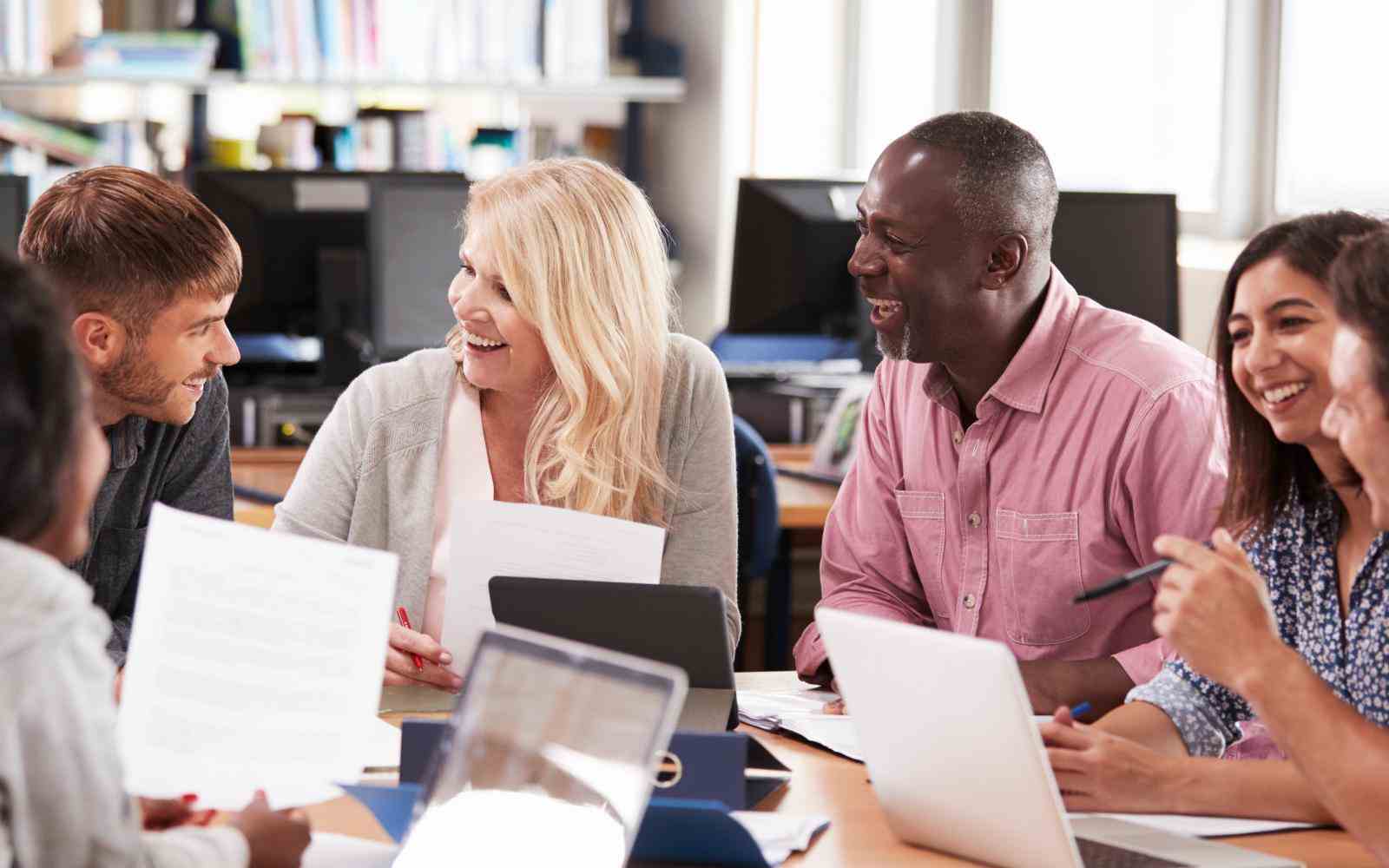 Grupo de pessoas estão conversando e sorrindo em torno de uma mesa em uma biblioteca. Algumas seguram folhas de papel, outras seguram canetas. Há tablets e computadores em cima da mesa