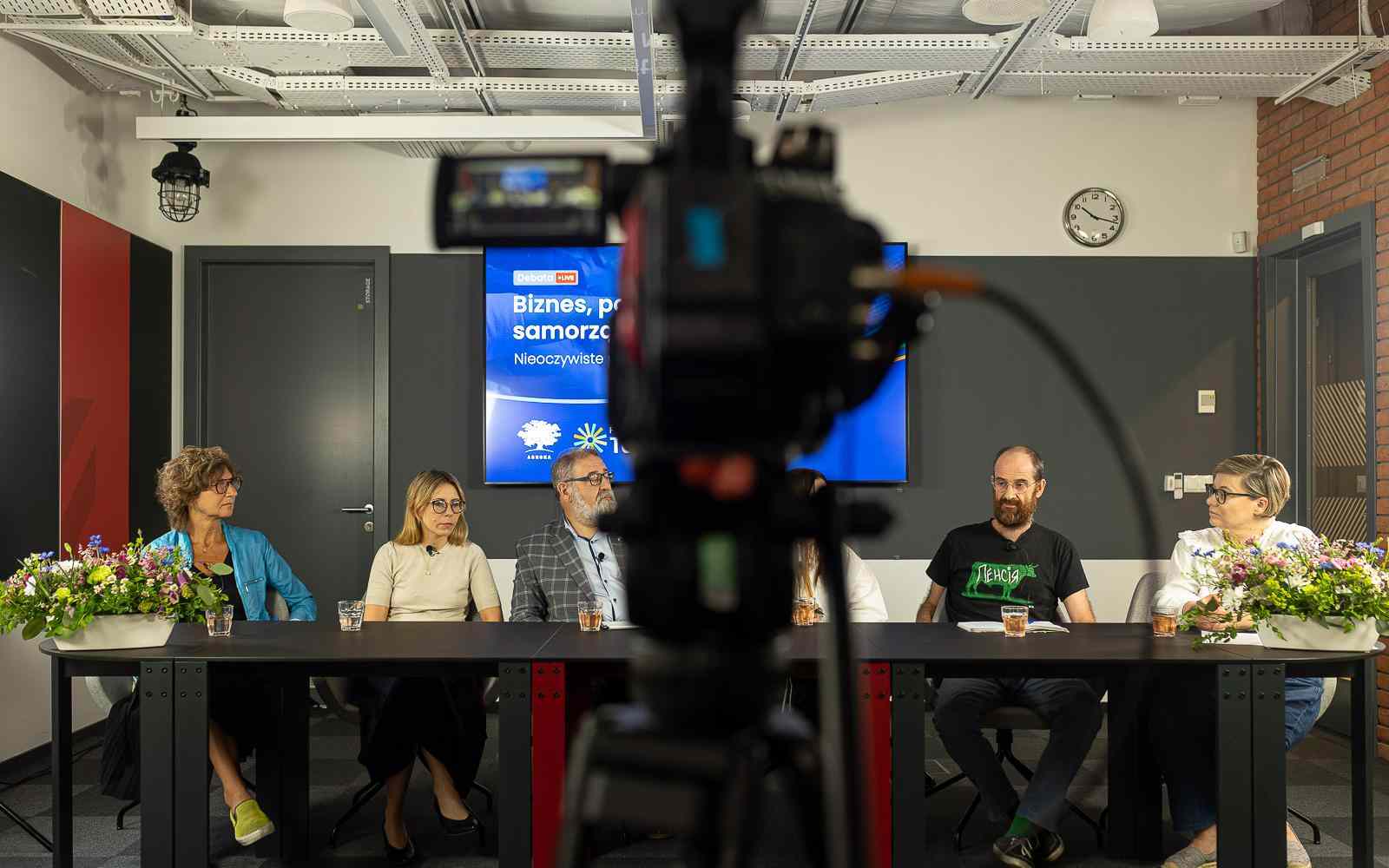 Zdjęcie przedstawia kamerę, która kręci debatę na środku kadru. Przy podłużnym stole siedzi 6 osób, które rozmawiają. Autorem zdjęcia jest Tomasz Kaczor.