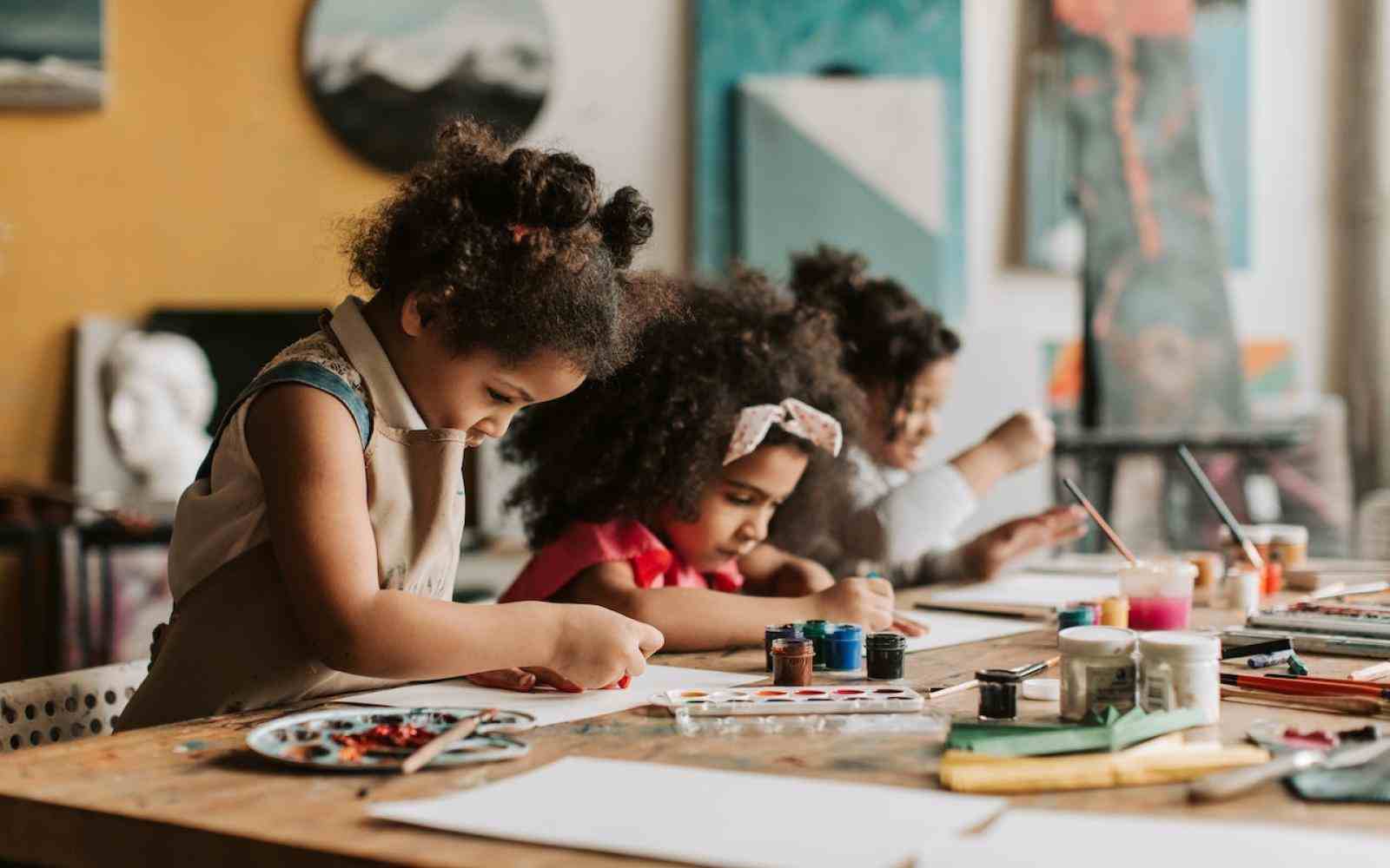 Duas crianças negras estão sentadas a uma mesa em que há tintas. Elas estão desenhando