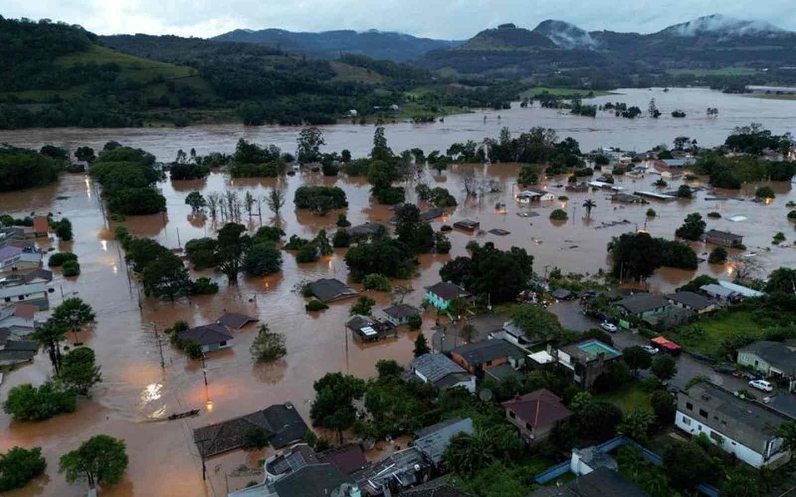Imagem aérea mostra uma cidade atingida por enchentes, com a água em tom amarronzado chegando até o telhado das casas e copa das árvores. Ao fundo, vê-se montanhas