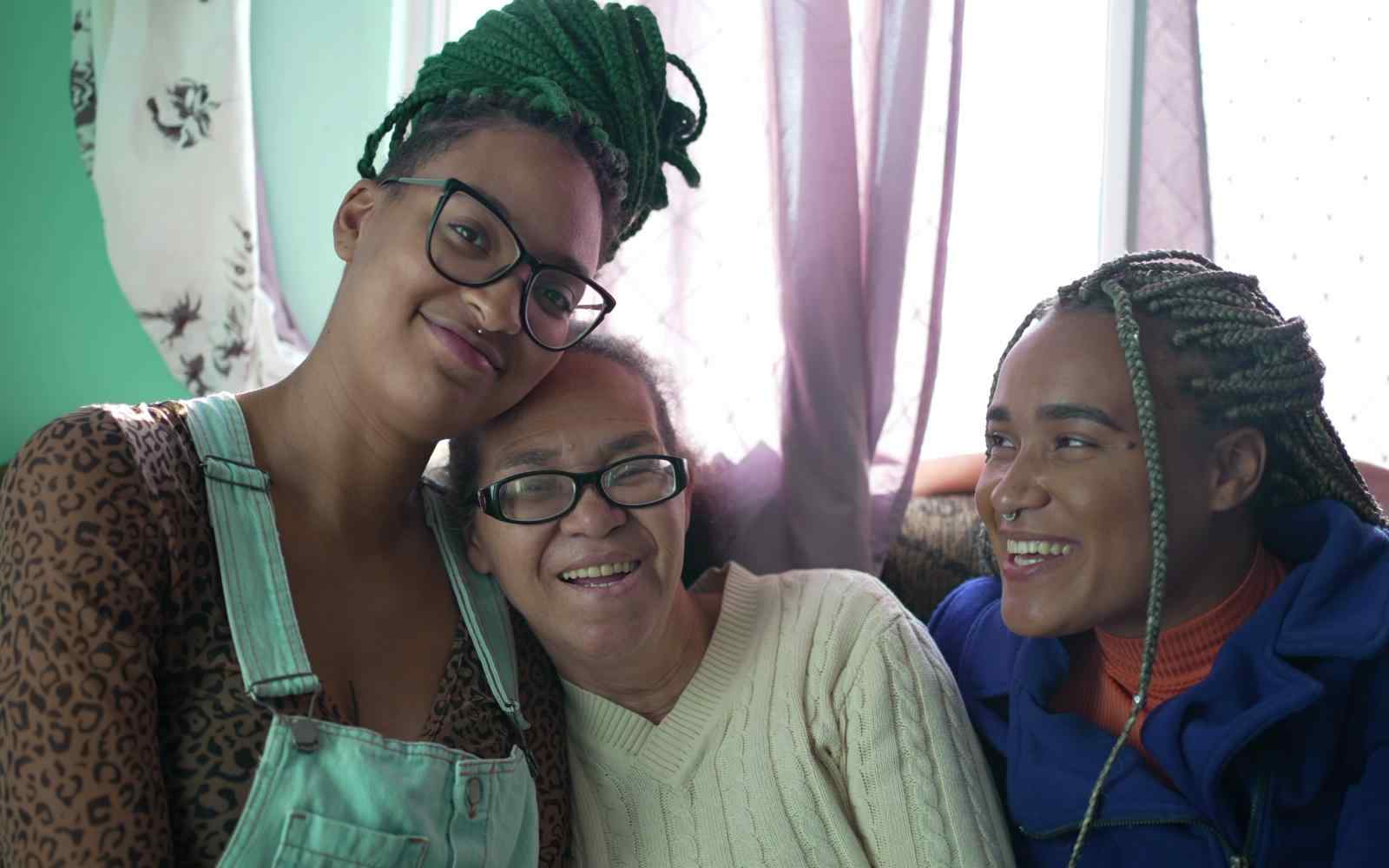 A imagem mostra três mulheres negras de diferentes gerações, sorrindo e sentadas em um sofá. No centro, há uma mulher mais velha, com cabelos grisalhos e óculos, que está sorrindo. Ela está cercada por duas pessoas mais jovens. A pessoa à esquerda usa óculos, tem tranças verdes e veste uma camisa estampada com uma jardineira jeans. A pessoa à direita, com tranças loiras, está vestida com uma jaqueta azul e também sorri