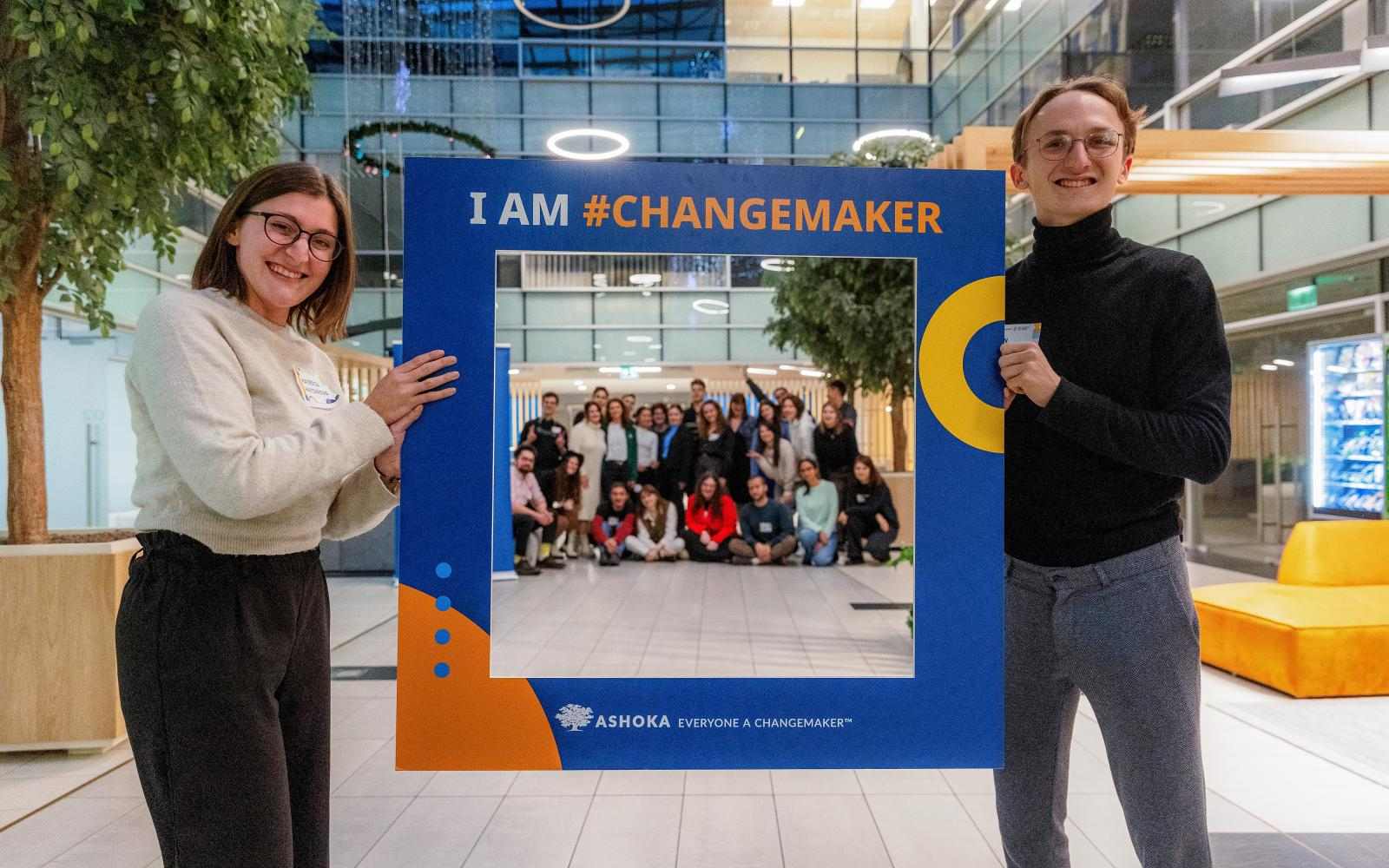 Two people holding a frame with caption "I AM #CHANGEMAKER", a group of people posing in the back in the middle of the frame