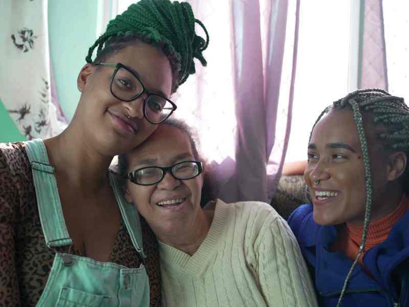 A imagem mostra três mulheres negras de diferentes gerações, sorrindo e sentadas em um sofá. No centro, há uma mulher mais velha, com cabelos grisalhos e óculos, que está sorrindo. Ela está cercada por duas pessoas mais jovens. A pessoa à esquerda usa óculos, tem tranças verdes e veste uma camisa estampada com uma jardineira jeans. A pessoa à direita, com tranças loiras, está vestida com uma jaqueta azul e também sorri