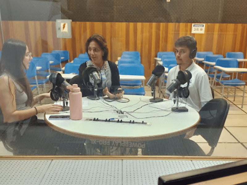 Três pessoas estão em um estúdio de gravação. Elas se sentam diante de uma mesa redonda de frente a microfones, e usam fones. Em primeiro plano, há um computador e uma mesa de áudio. Um vidro parece separar o grupo da mesa de áudio