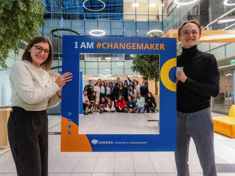 Two people holding a frame with caption "I AM #CHANGEMAKER", a group of people posing in the back in the middle of the frame