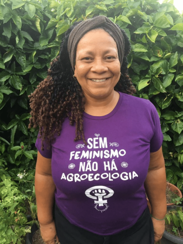 Beth Cardoso is a black woman. She has dark curly hair and is wearing a cross hair band and a purple T-shirt with the saying "Without feminism there isn't agroecology" and the feminist symbol